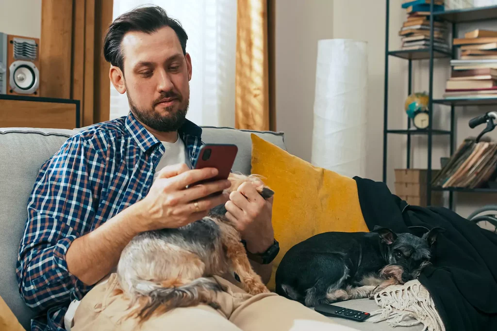 Disabled man on couch with two dogs using mobile phone to contact Emerging Supports